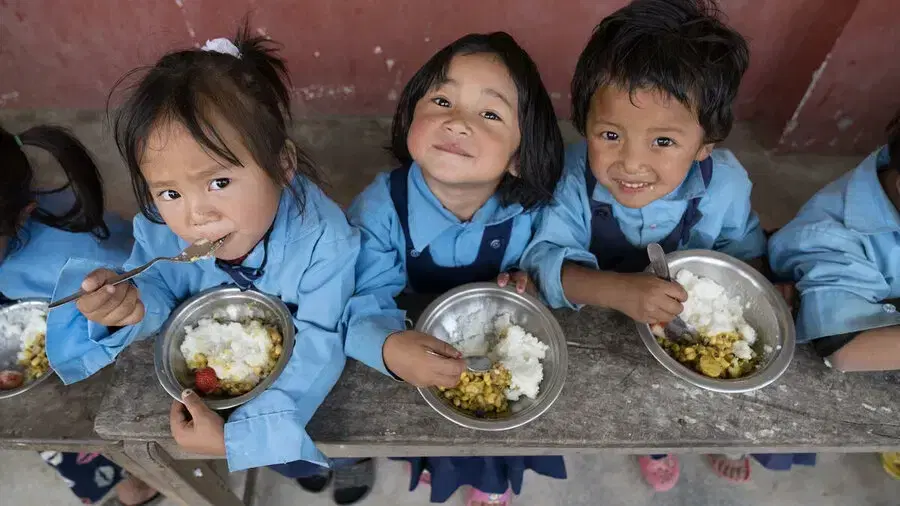 Three smiling young children in blue school uniforms eating rice and lentils from steel bowls