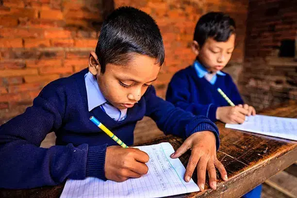 Two young students in navy jumpers writing in their exercise books at a wooden desk